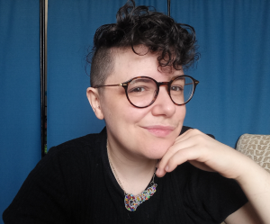 A white human with round tortoise shell glasses and short dark curly hair on top with shaved sides. They are wearing a black short sleeved t-shirt and a colourful beaded necklace which spells out the word queer. They are resting their chin on one hand, with a cheeky smile. The background is a blue room screen.