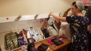 A white person, Vox, is looking at a dressing room mirror, and seems to be screwing in a light bulb. They are dressed in a blue dress with a floral pattern and are wearing a headscarf in a vintage style. On the table in front of the mirror are props and makeup. In the reflection you can see Vox, and their smile. You can also see in the reflection a number of colourful costumes hung up on the wall behind Vox. 