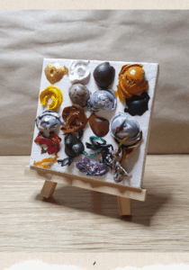 A bigger version of the art described on the previous page. More details of the art: The domes of paint have various cracks in them, some have a hold in the centre. The colours of the paint are: gold, white, black, orange, yellow, brown, grey mixed with white and some red, green, purple, and a rainbow bit. Description from previous page: Image shows a square canvas on a small easel placed on a desk. The canvas has 4 columns and 5 rows of domes of dried paint. The paint get decreasingly circular towards the bottom of the canvas.